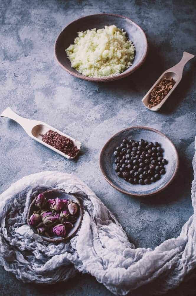 Five ceramic bowls and scoops containing dried rosebuds, juniper berries, brown seeds, cloves, and a light green powder are arranged on a gray textured surface with a white cloth.