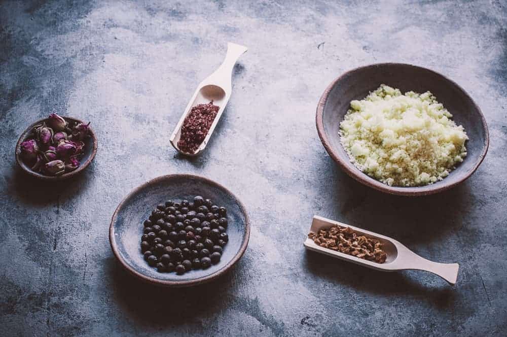 A flat lay of bowls and scoops containing dried rosebuds, black berries, brown spice, pink salt, and a bowl of grated or crumbled yellow ingredient on a gray surface.