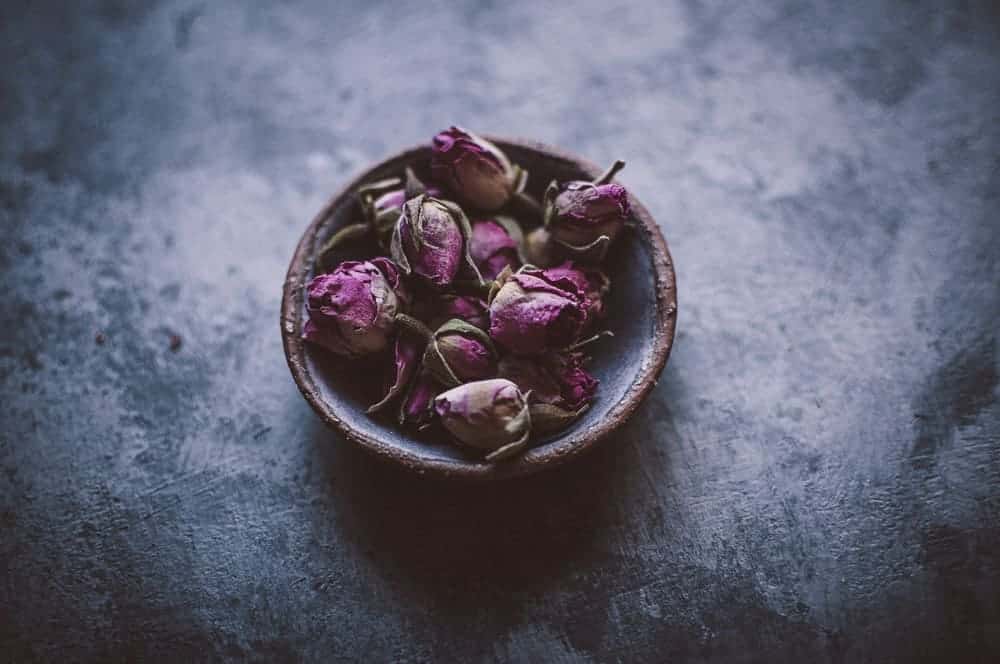 A small ceramic bowl filled with dried pink rosebuds sits on a textured dark surface.