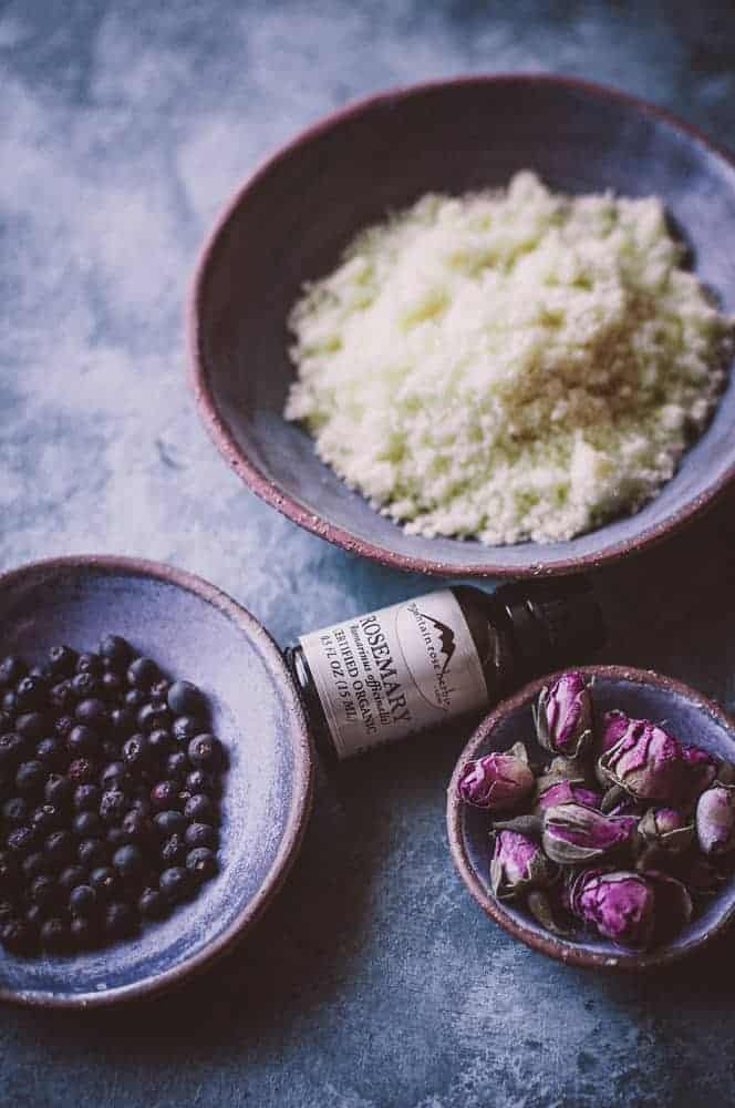 Three ceramic bowls containing dried blueberries, pale green granules, and dried rosebuds are arranged beside a small glass bottle labeled “Rosemary Art.”.
