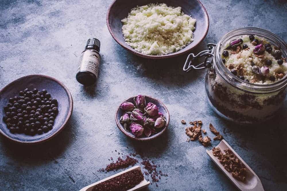 A jar and bowls containing dried rosebuds, dark berries, a bottle of oil, and a coarse mixture are arranged on a textured surface with small wooden scoops.