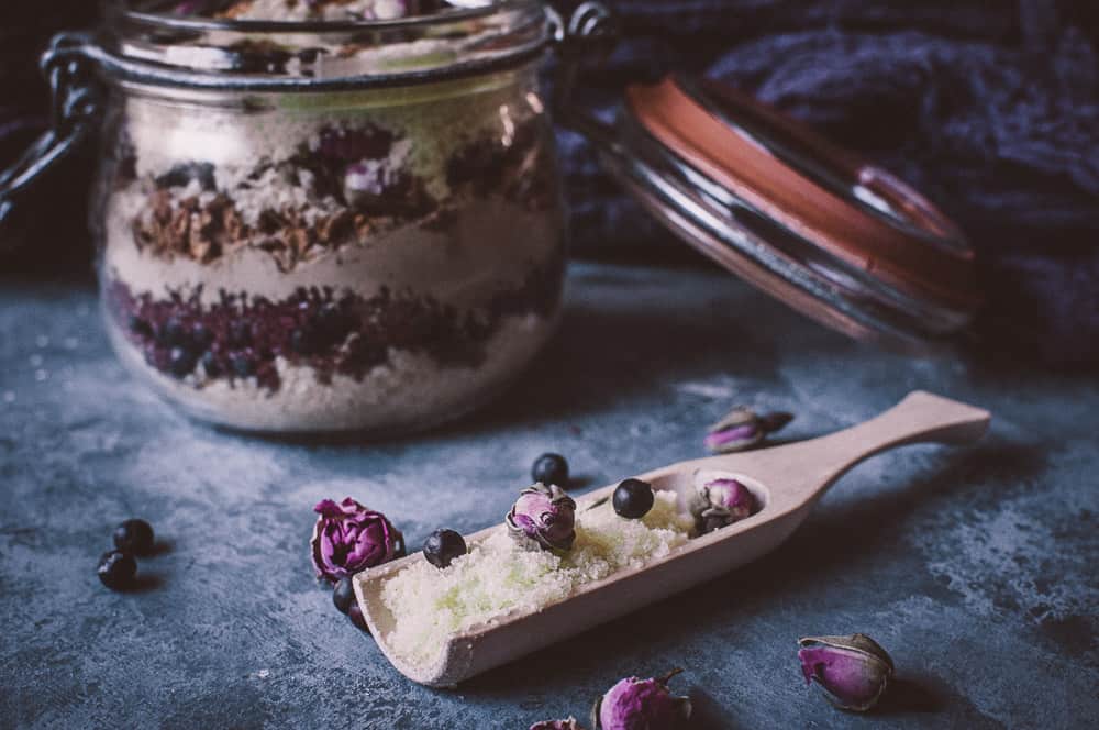 A glass jar of layered bath salts with dried flowers and berries sits next to an open wooden scoop containing bath salts, roses, and berries on a textured surface.