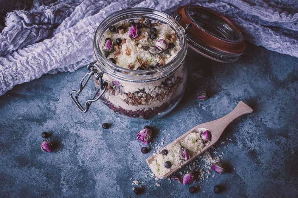 A glass jar filled with layered ingredients, topped with dried rosebuds and berries, sits on a textured surface next to a wooden scoop holding a similar mixture.