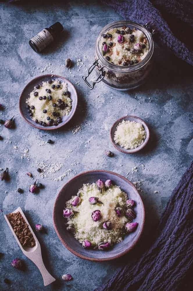A flat lay of bowls and a jar containing a homemade bath scrub with dried flowers and berries, surrounded by scattered ingredients on a textured surface.