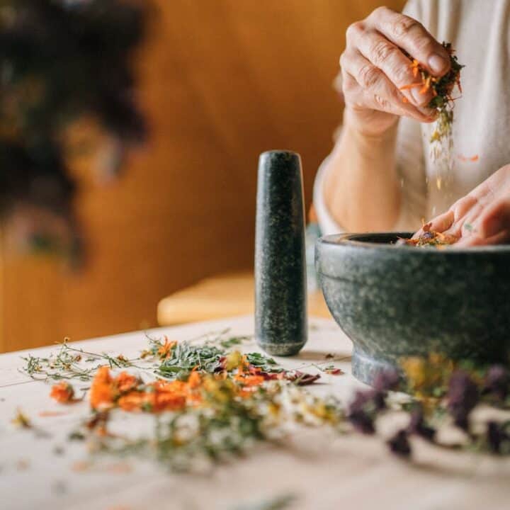 Person grinding dried flowers and herbs in a stone mortar and pestle on a light wooden table, with scattered plant material around.