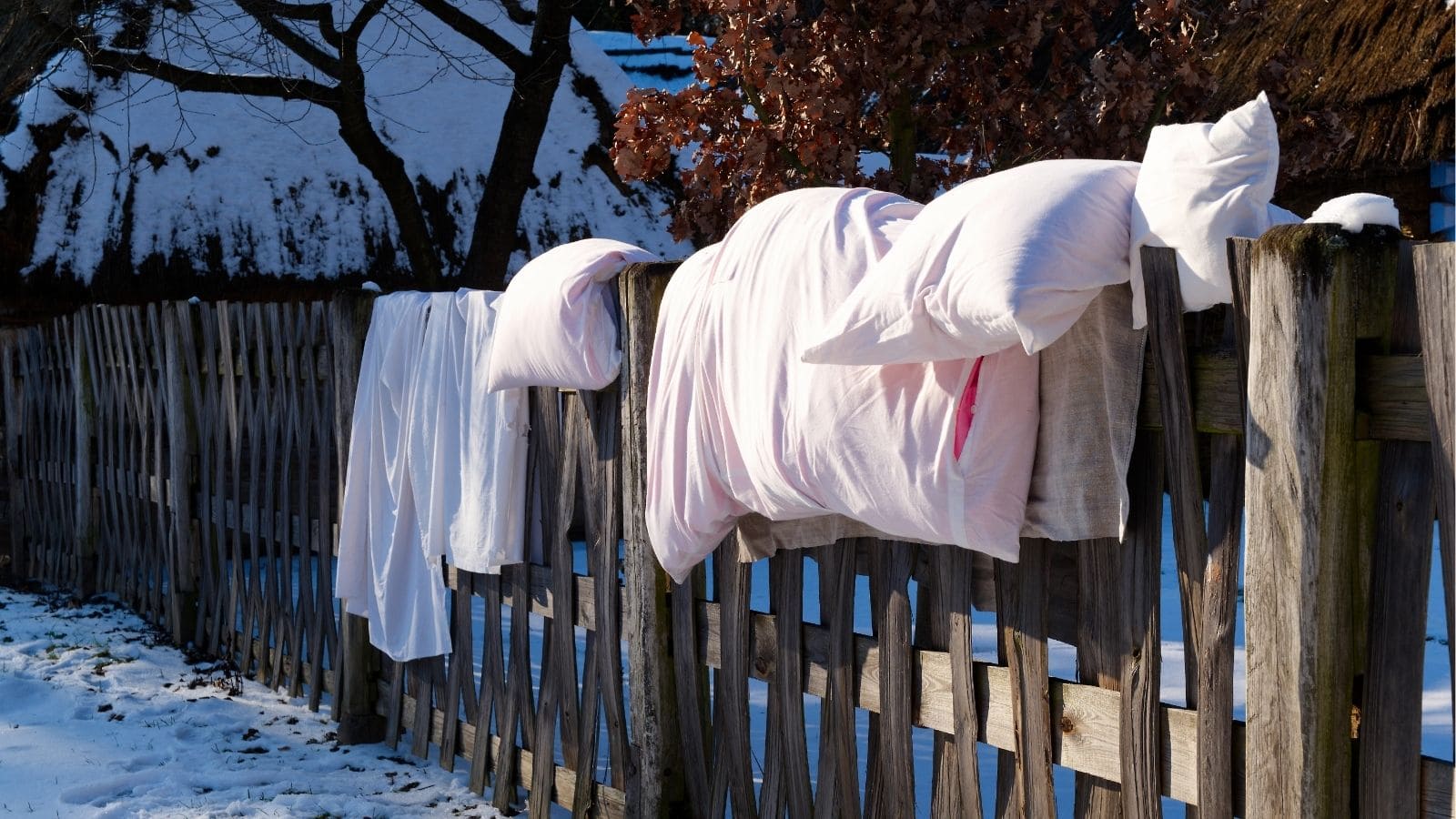 Pillows and bed linens are draped over a wooden fence to air out, with snow on the ground and trees in the background.