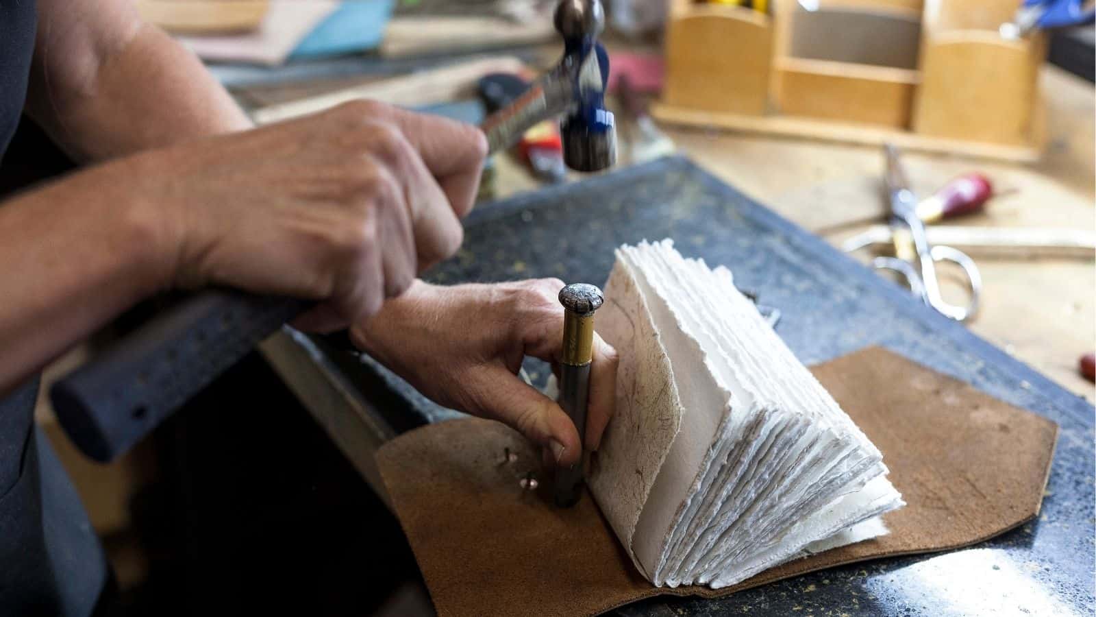 A person uses a hammer and metal tool to punch holes in a stack of rough-edged paper sheets on a workbench.
