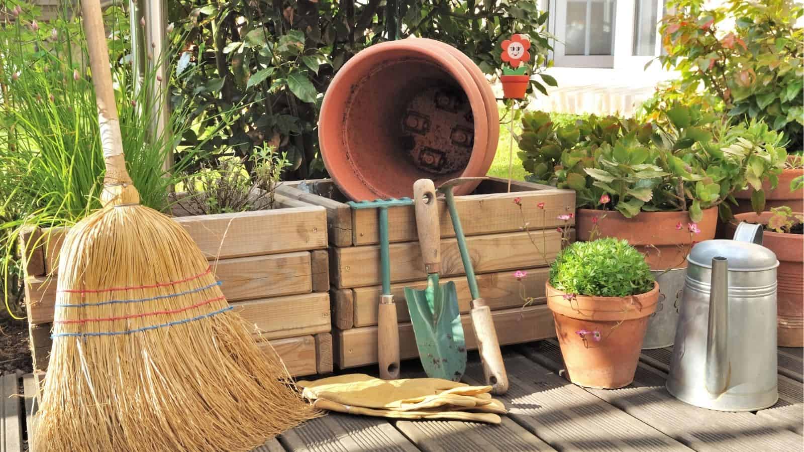 A broom, gloves, trowel, and small shovel are arranged with potted plants and a watering can on a garden deck in front of wooden raised beds.