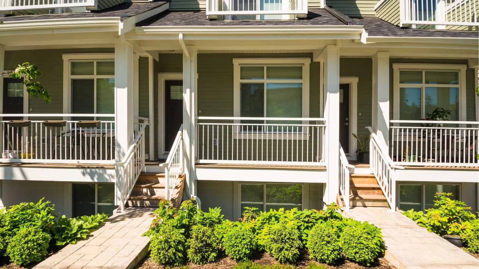 Exterior view of a modern townhouse with two front porches, white railings, and green siding, featuring stairs and landscaping with bushes.