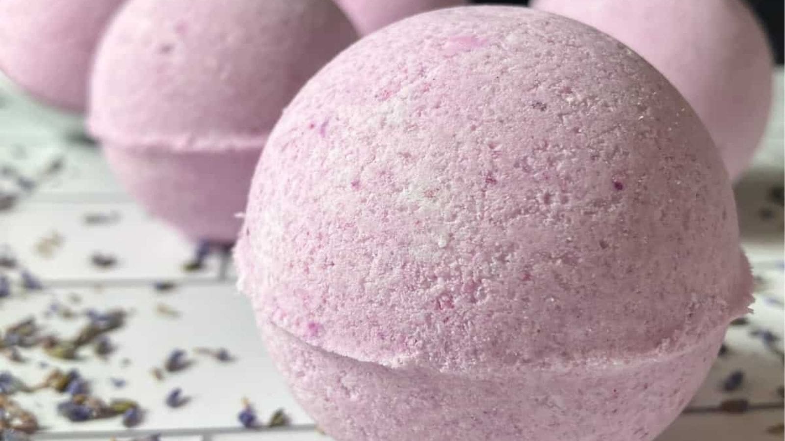 Close-up of round, pink bath bombs with a textured surface, placed on a white tiled surface with dried lavender scattered around.