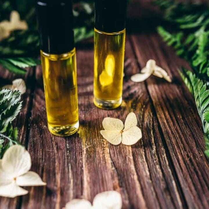 Two glass bottles filled with yellow oil are placed on a wooden surface, surrounded by green fern leaves and white dried flowers.