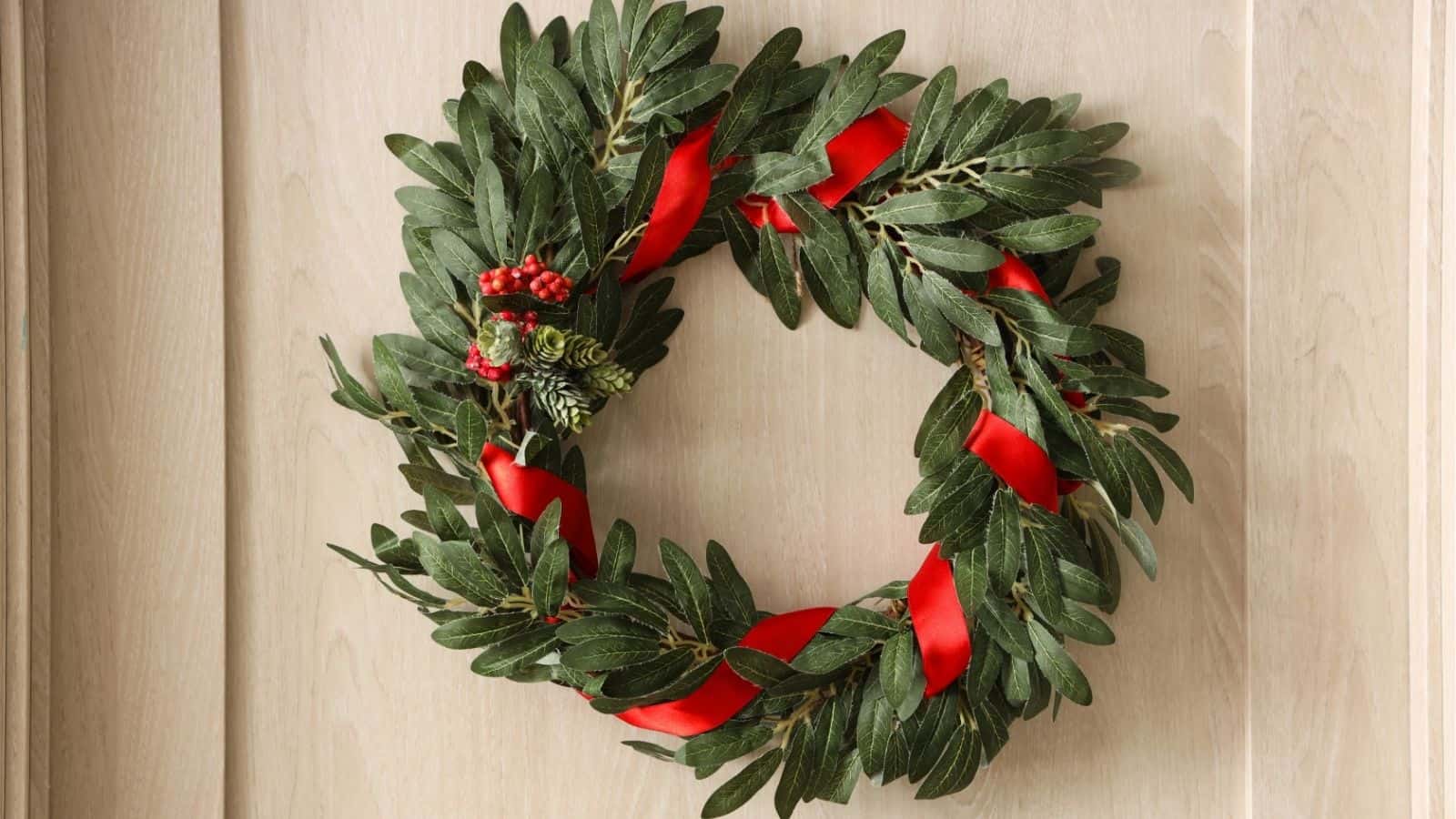 A green wreath with red berries, pine sprigs, and a red ribbon is hung on a light wooden door.