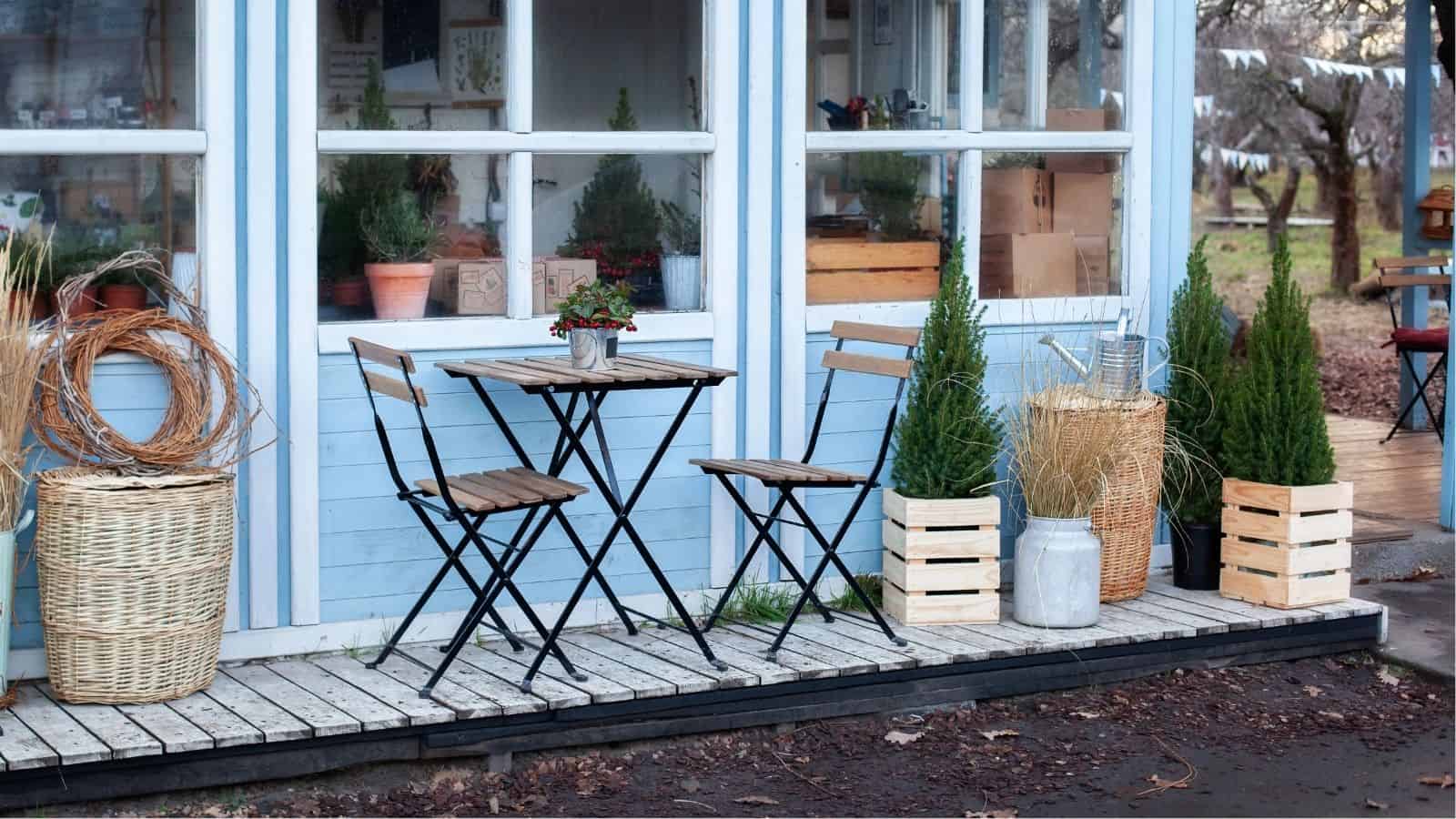 A small outdoor patio features a wooden table with two chairs, potted plants, baskets, wooden crates, and a glass door leading inside.