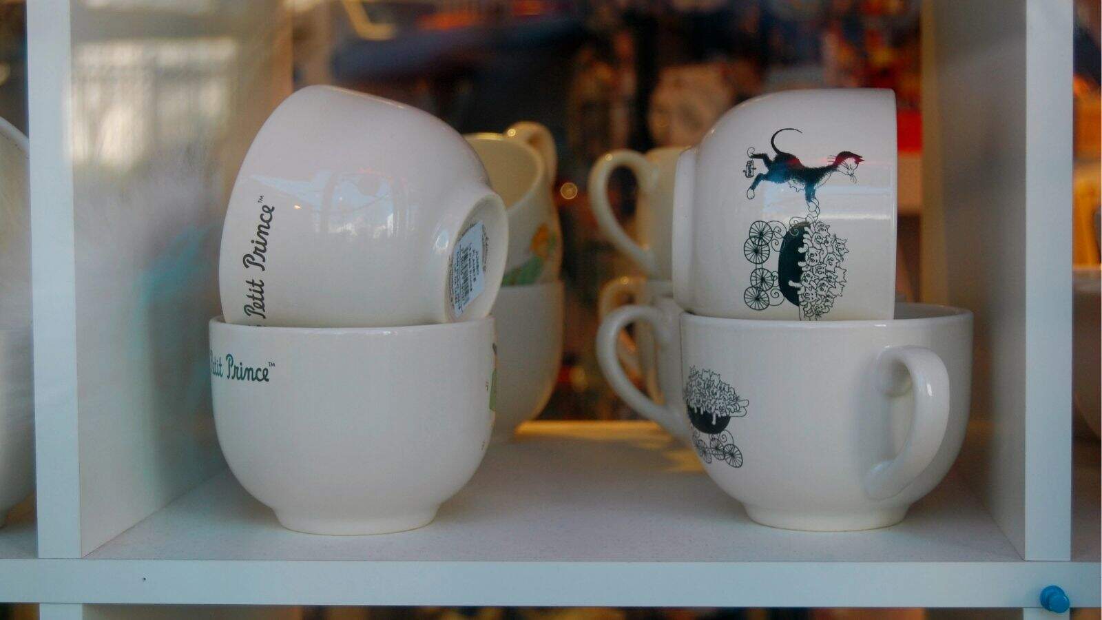 Four white ceramic mugs with whimsical designs and the words "Butt Prince" are stacked on a shelf, with other blurred mugs and items in the background.