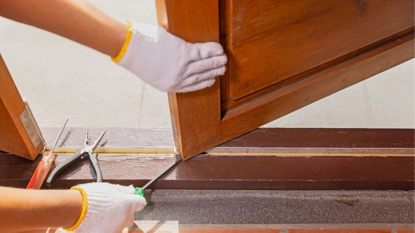 Person wearing white gloves uses a screwdriver to install or repair a wooden door, with pliers and a screwdriver lying nearby on the door frame.