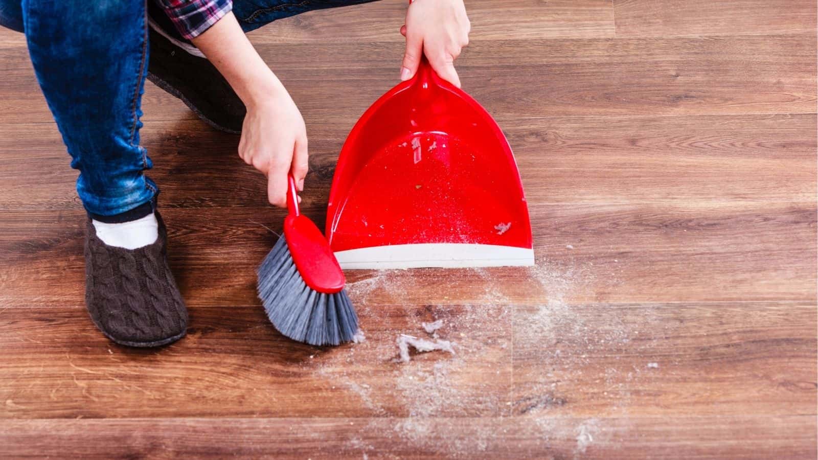 Person sweeping dust and debris from a wooden floor into a red dustpan using a handheld brush.