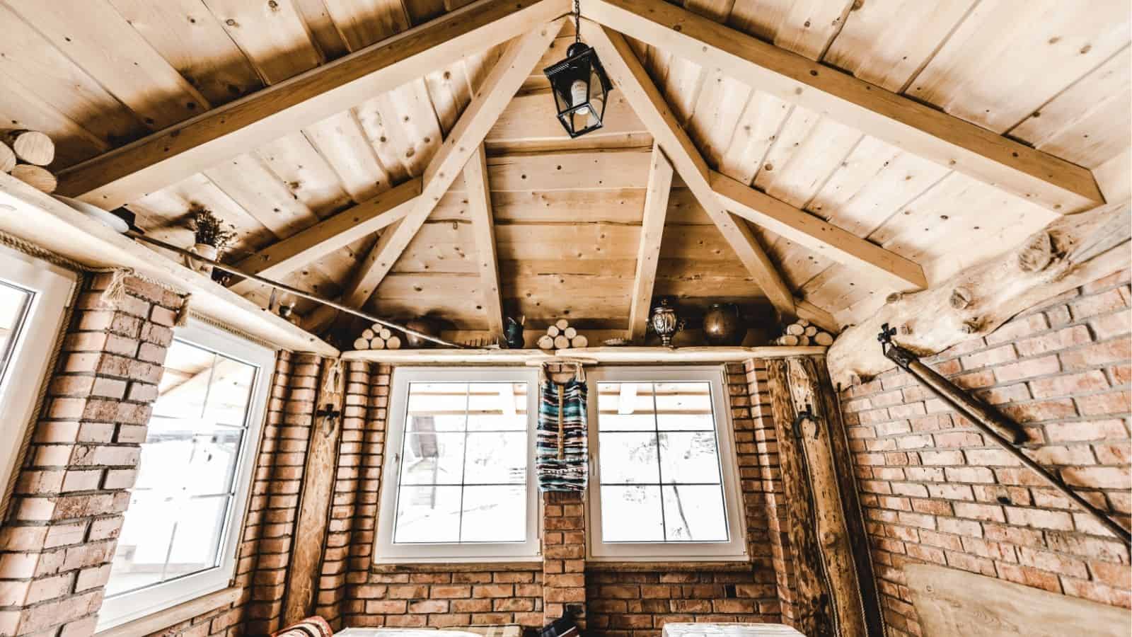 Rustic wooden ceiling and brick walls with two windows, stacked firewood, and a lantern in a cozy cabin interior.