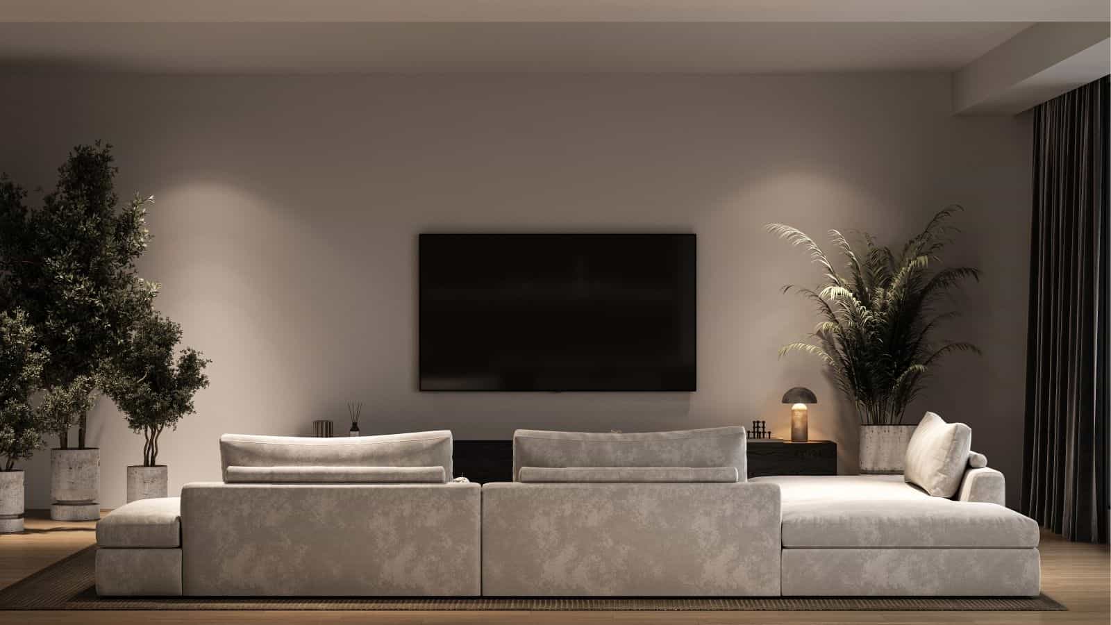 Modern living room with a large gray sectional sofa facing a wall-mounted flat-screen TV, flanked by tall potted plants and a sideboard with a lamp and decor.