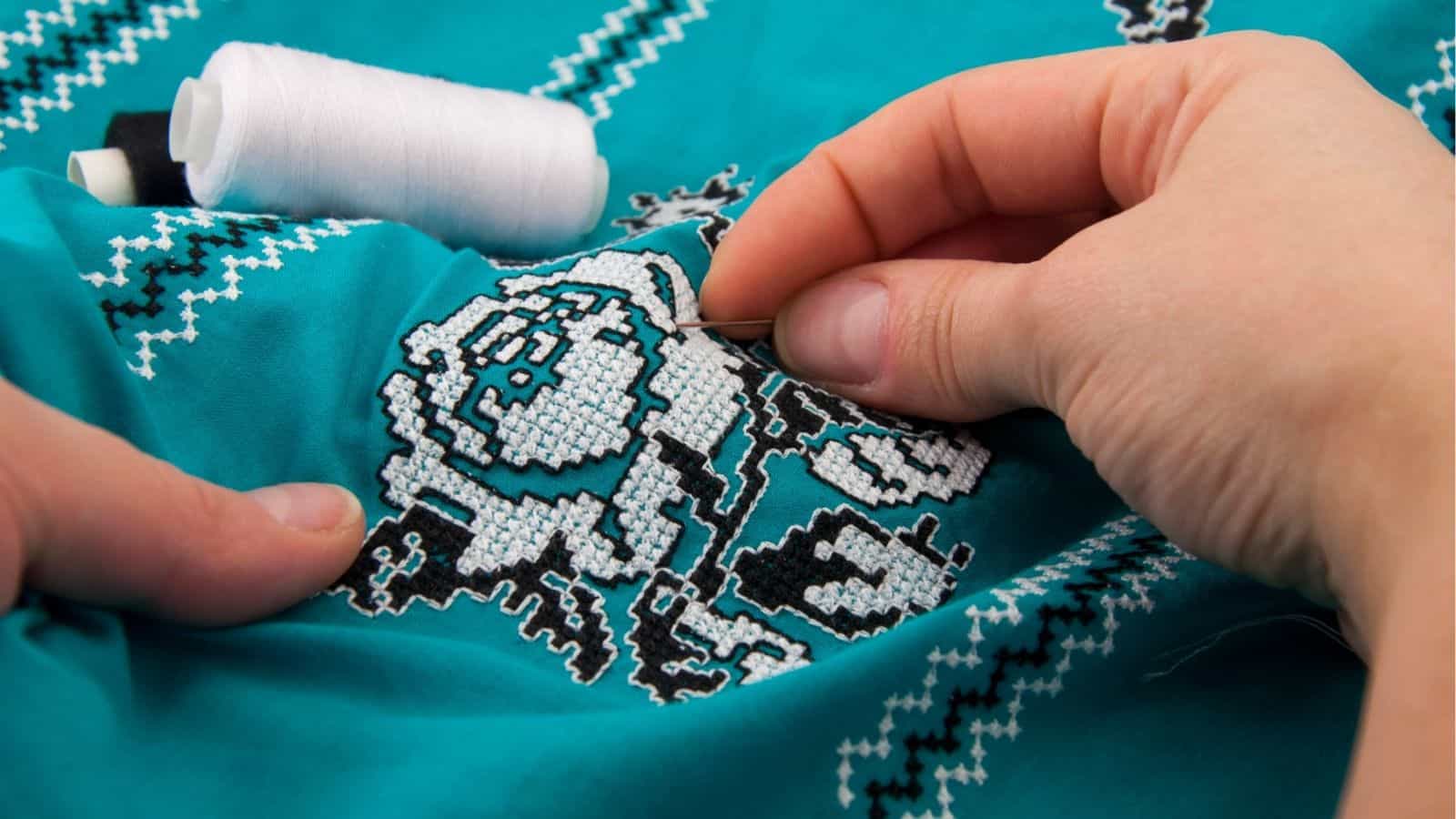 Close-up of hands embroidering a floral pattern with white and black thread on turquoise fabric, with a spool of white thread nearby.