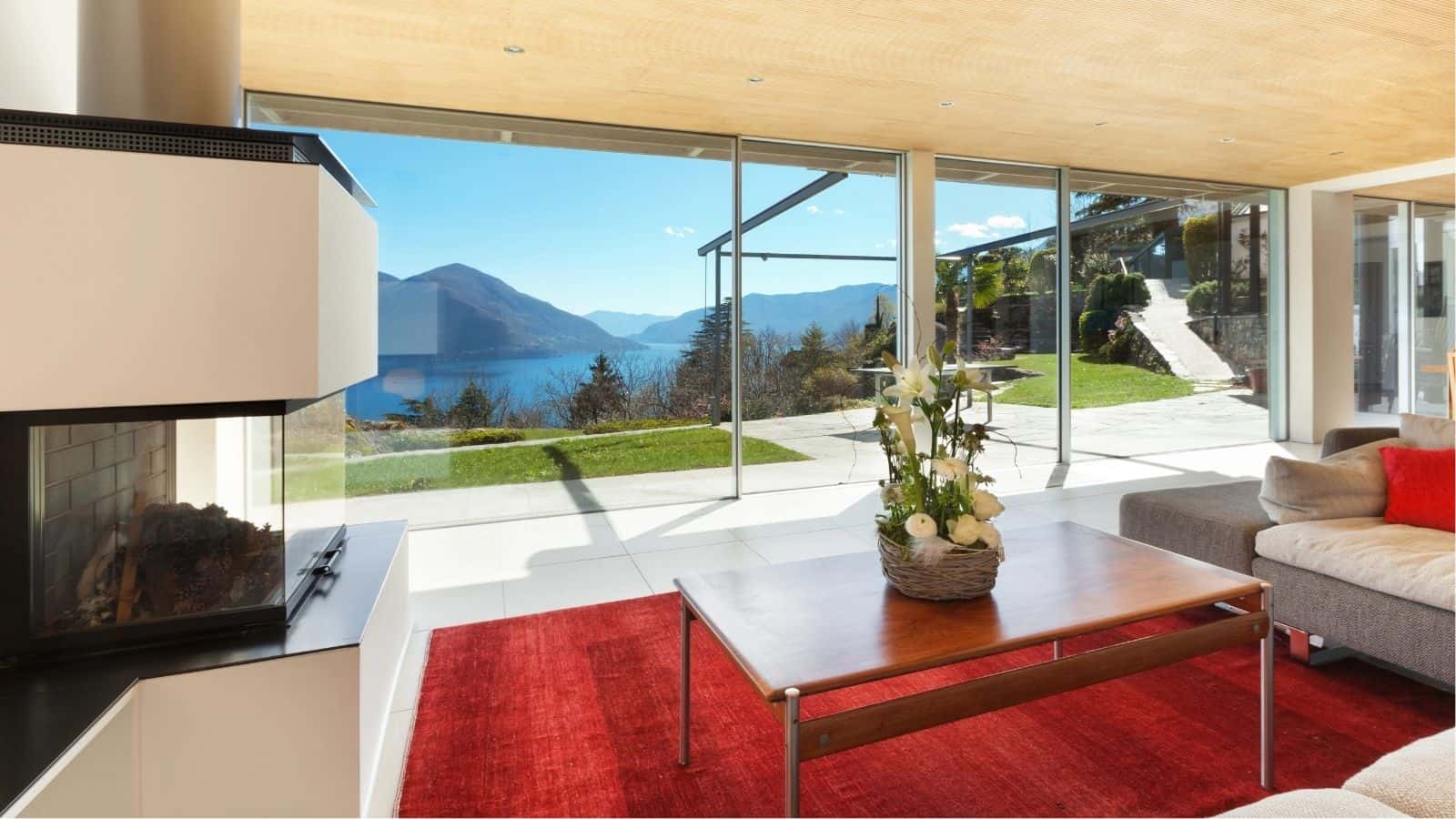Modern living room with large glass windows offering a view of mountains, a lake, and a patio; red rug and wooden table with a floral arrangement in the foreground.