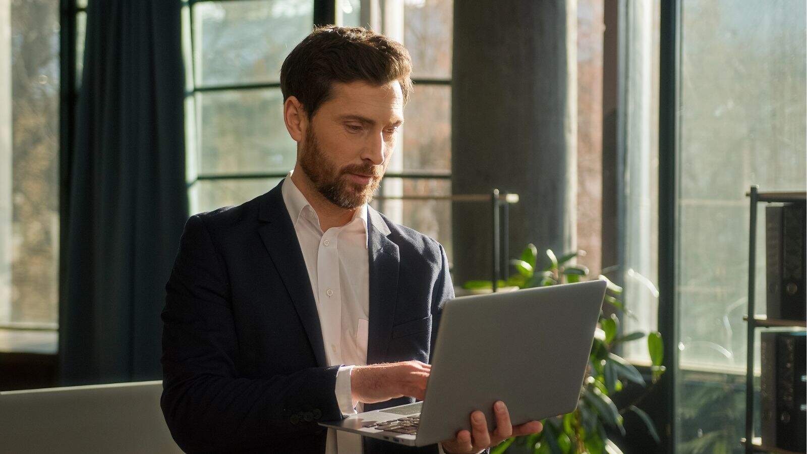 A man in a suit stands indoors, using a laptop, with sunlight streaming through large windows behind him.