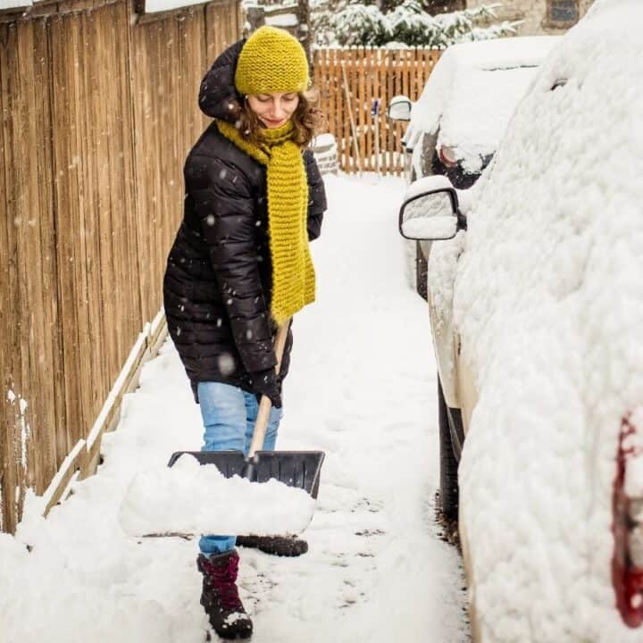 A person wearing a winter coat, hat, and scarf shovels snow beside a car parked next to a wooden fence.