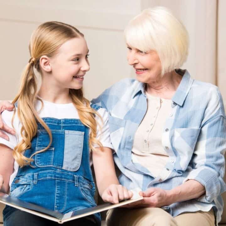 An older woman and a young girl sit on a couch smiling at each other while looking at an open book in the girl's lap.