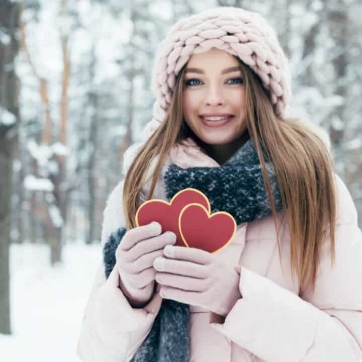 A young woman in winter clothing holds two red heart-shaped cookies while standing in a snowy forest.