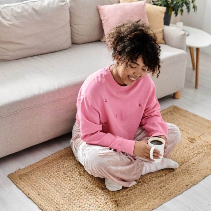 A person sits on a woven rug in front of a sofa, holding a cup of coffee and smiling, in a bright living room with light-colored decor.