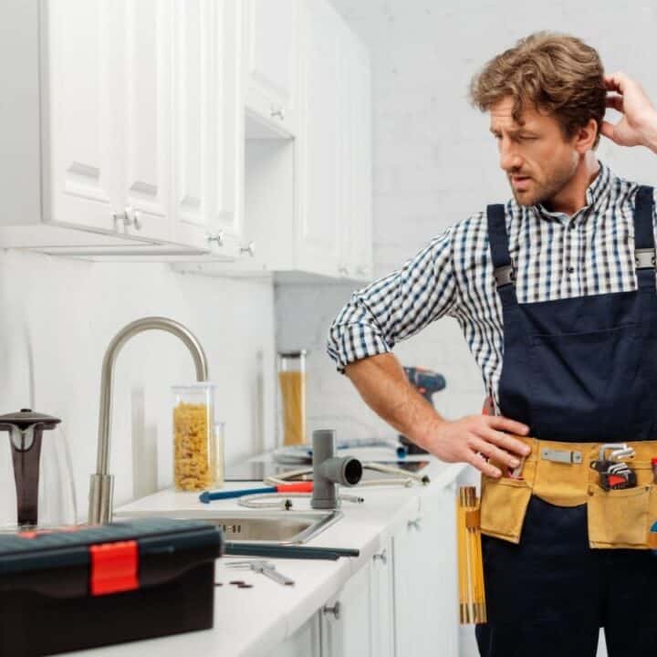 A man in work overalls stands in a kitchen, scratching his head in confusion while looking at pipes and tools on the counter.