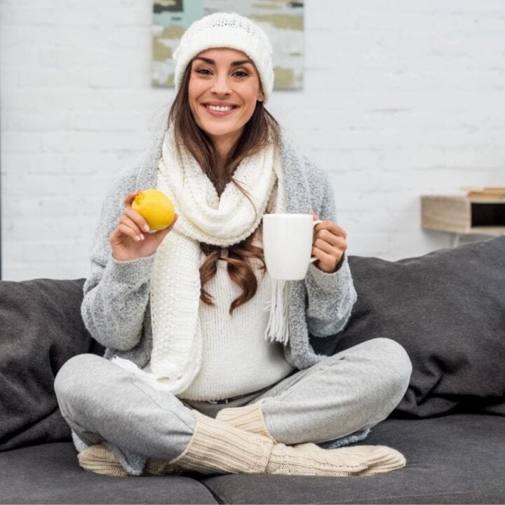 A woman in winter clothing sits cross-legged on a sofa, holding a yellow apple in one hand and a white mug in the other, and smiling at the camera.