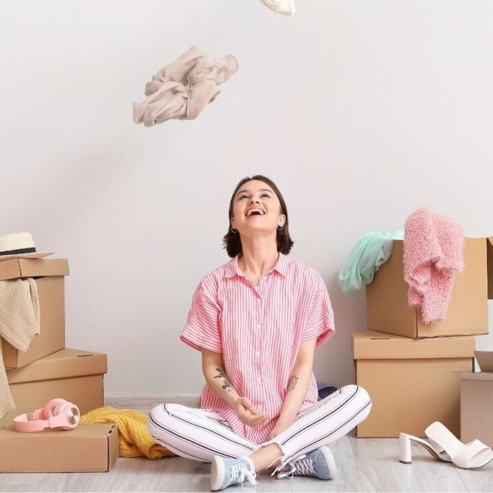A woman sits on the floor surrounded by cardboard boxes and clothes, tossing an item of clothing in the air.