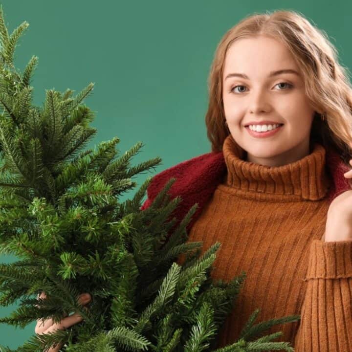 A young woman in a brown sweater holds a small Christmas tree against a green background, smiling at the camera.