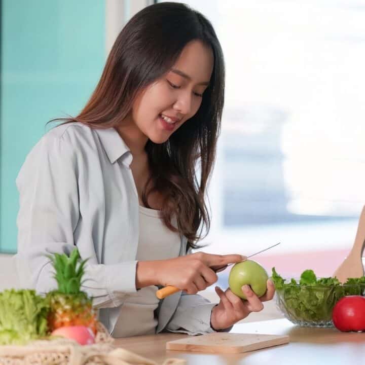 A woman slices a green apple at a kitchen counter with various fruits, vegetables, and a recycling bin nearby.