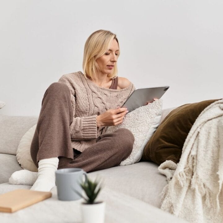 A woman sits on a sofa with a blanket and pillows, holding a tablet. A mug, book, and woven basket are nearby.