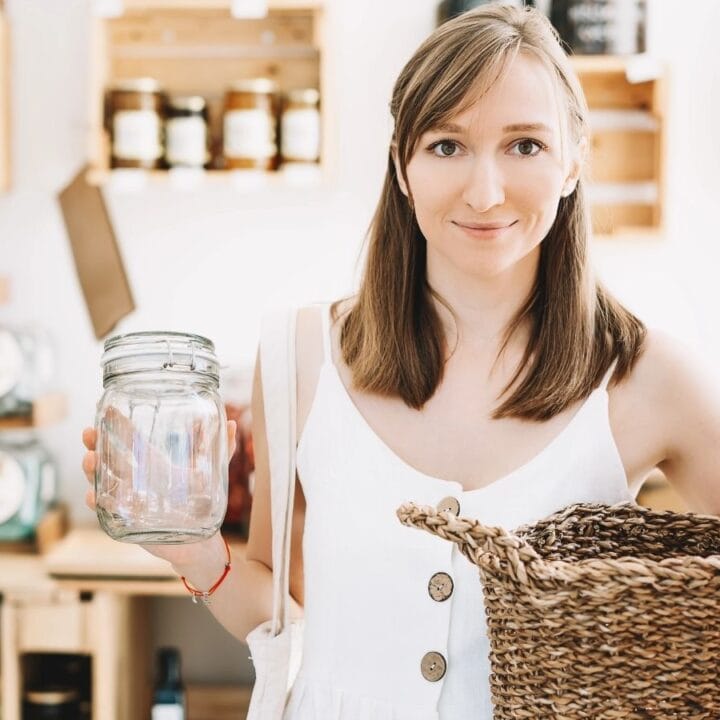 A woman in a white dress holds a glass jar and a woven basket while shopping in a store with jars and wooden shelves in the background.