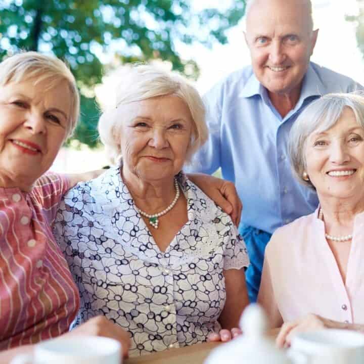 Four older adults, three women and one man, sit together at an outdoor table, smiling at the camera with trees and sunlight in the background.