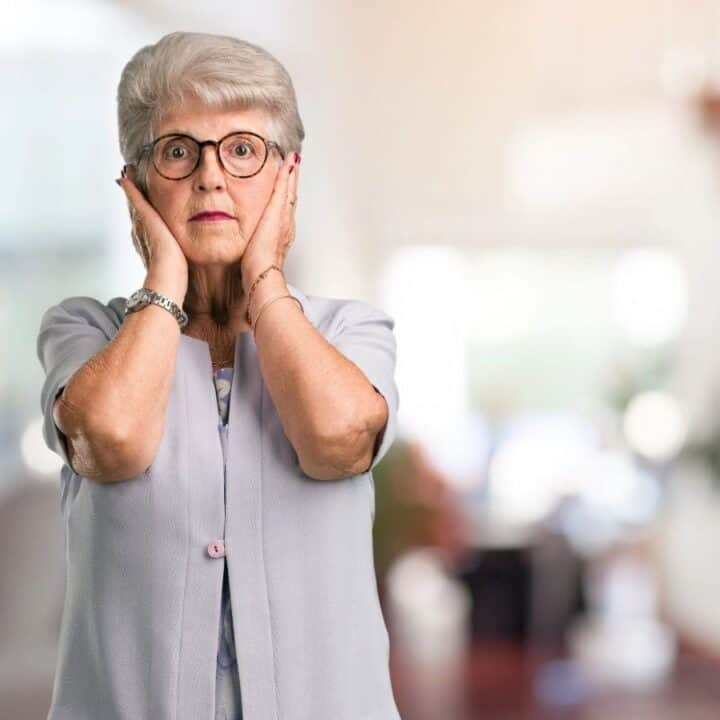 An elderly woman with gray hair and glasses holds her hands to her face, looking surprised, standing indoors with a blurred background.