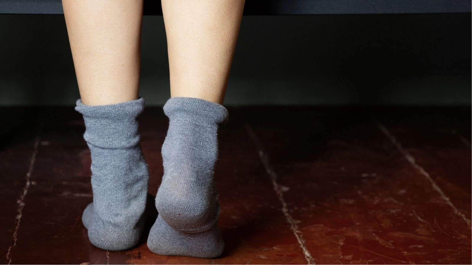 Close-up of two feet wearing gray socks standing on a reddish-brown wooden floor.