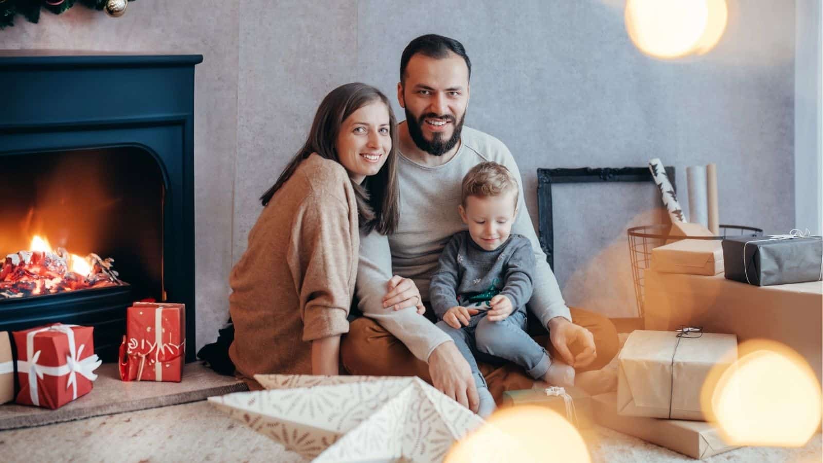 A family of three sits on the floor near a fireplace and wrapped gifts, smiling and playing together in a cozy living room.