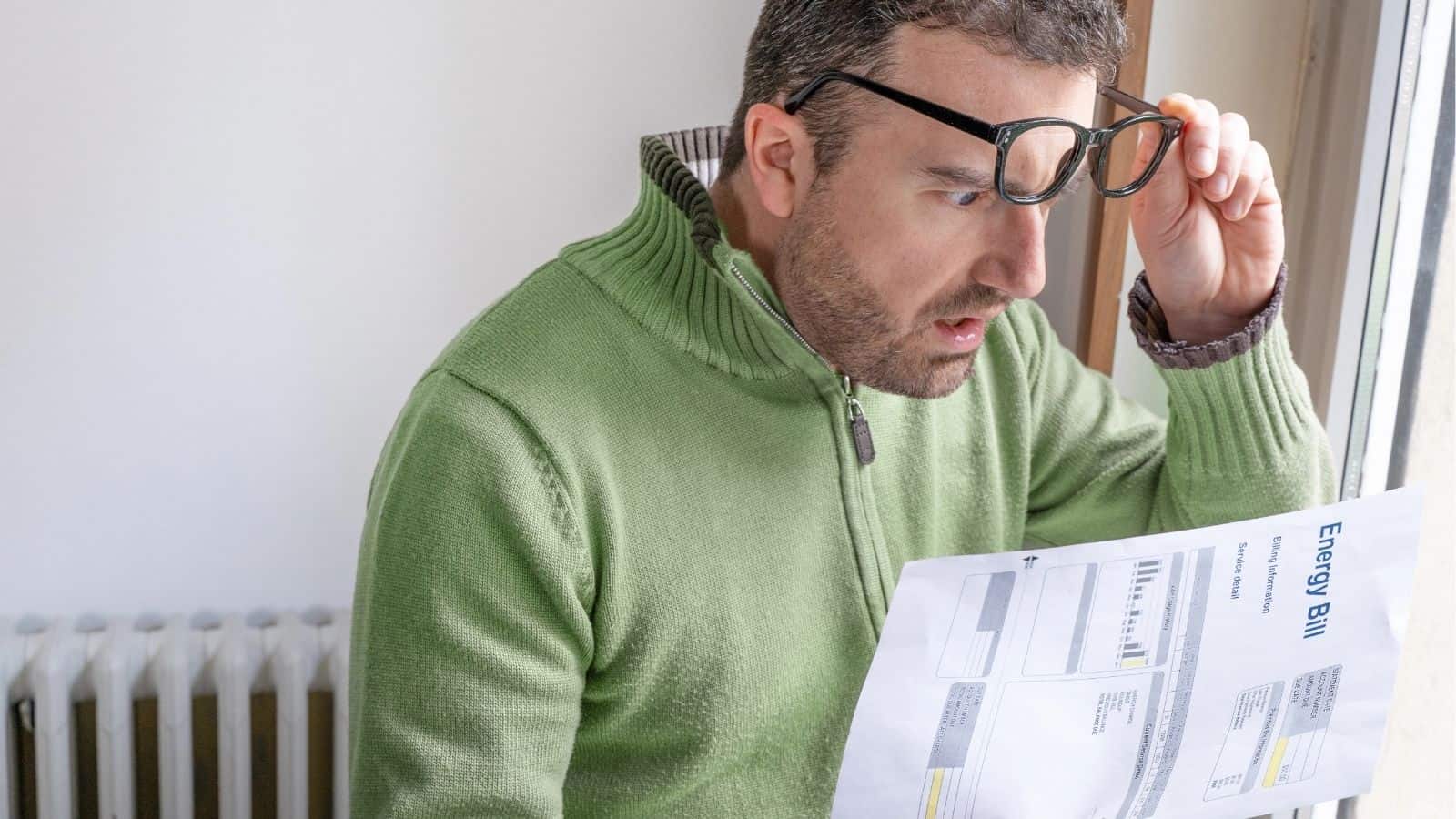 A man in a green sweater looks surprised while holding and reading an energy bill.