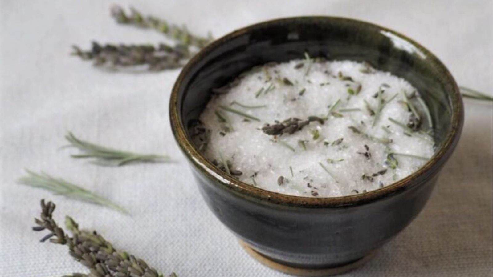 A ceramic bowl filled with coarse salt mixed with dried lavender sits on a light-colored fabric, with sprigs of lavender scattered nearby.
