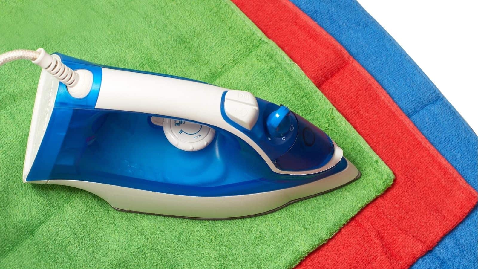 A blue and white electric iron placed on a green towel, with red and blue towels stacked underneath, on a white background.