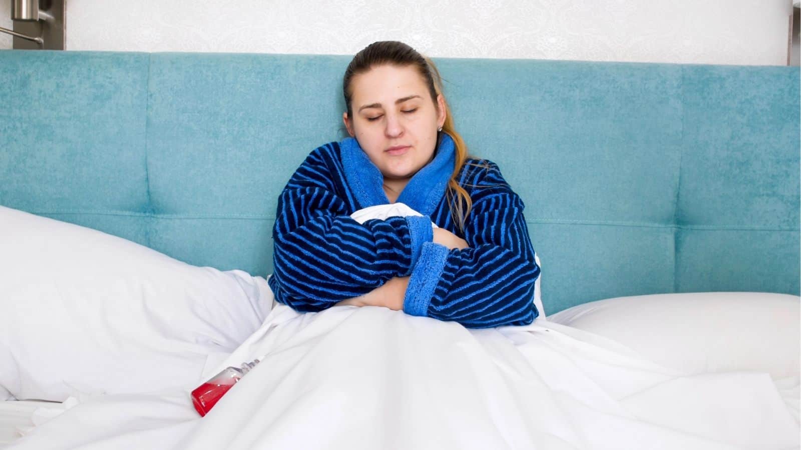 A woman in a blue robe sits up in bed with her eyes closed, arms crossed over her chest, and a medicine bottle on the blanket.