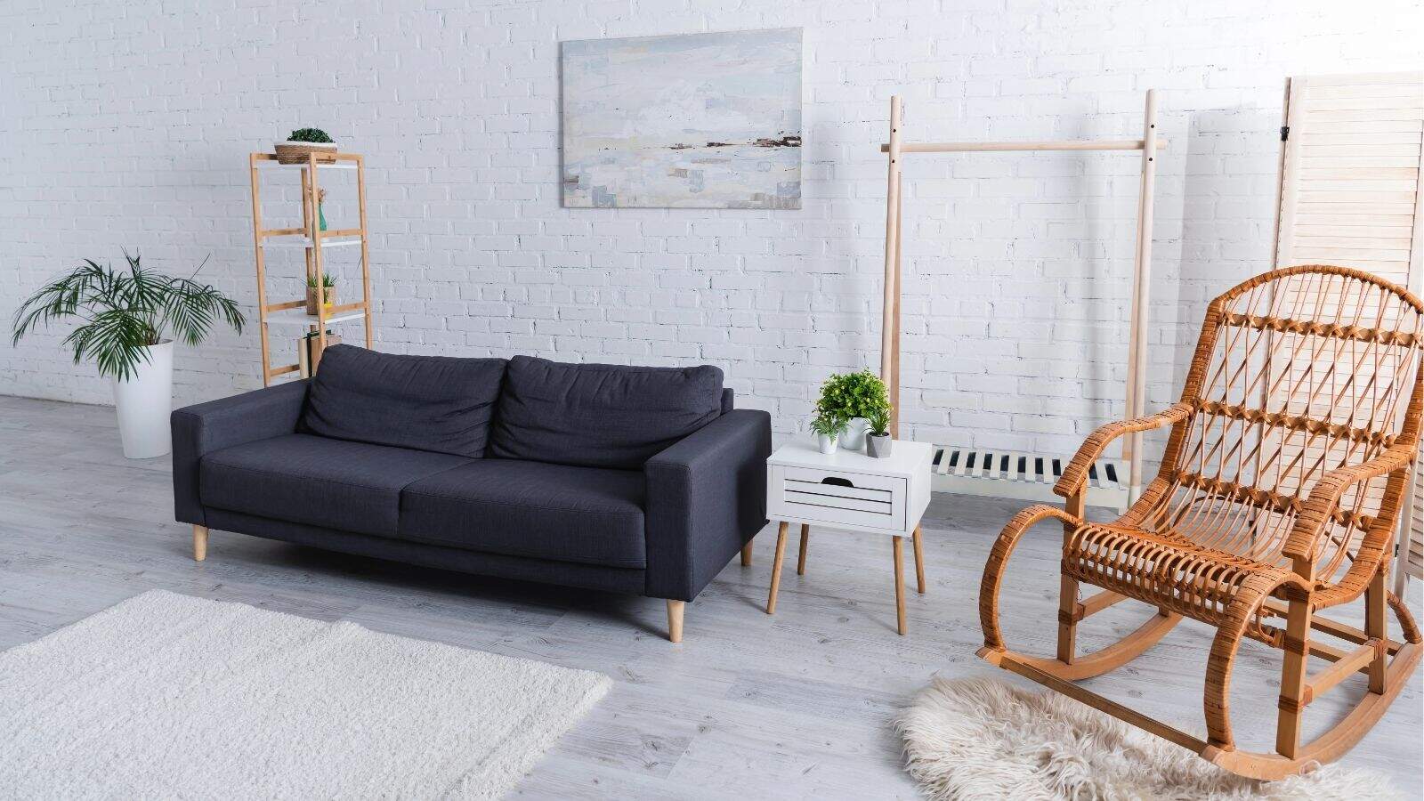 Modern living room with a dark gray sofa, wicker rocking chair, small white table, potted plants, and wall art against a white brick wall.