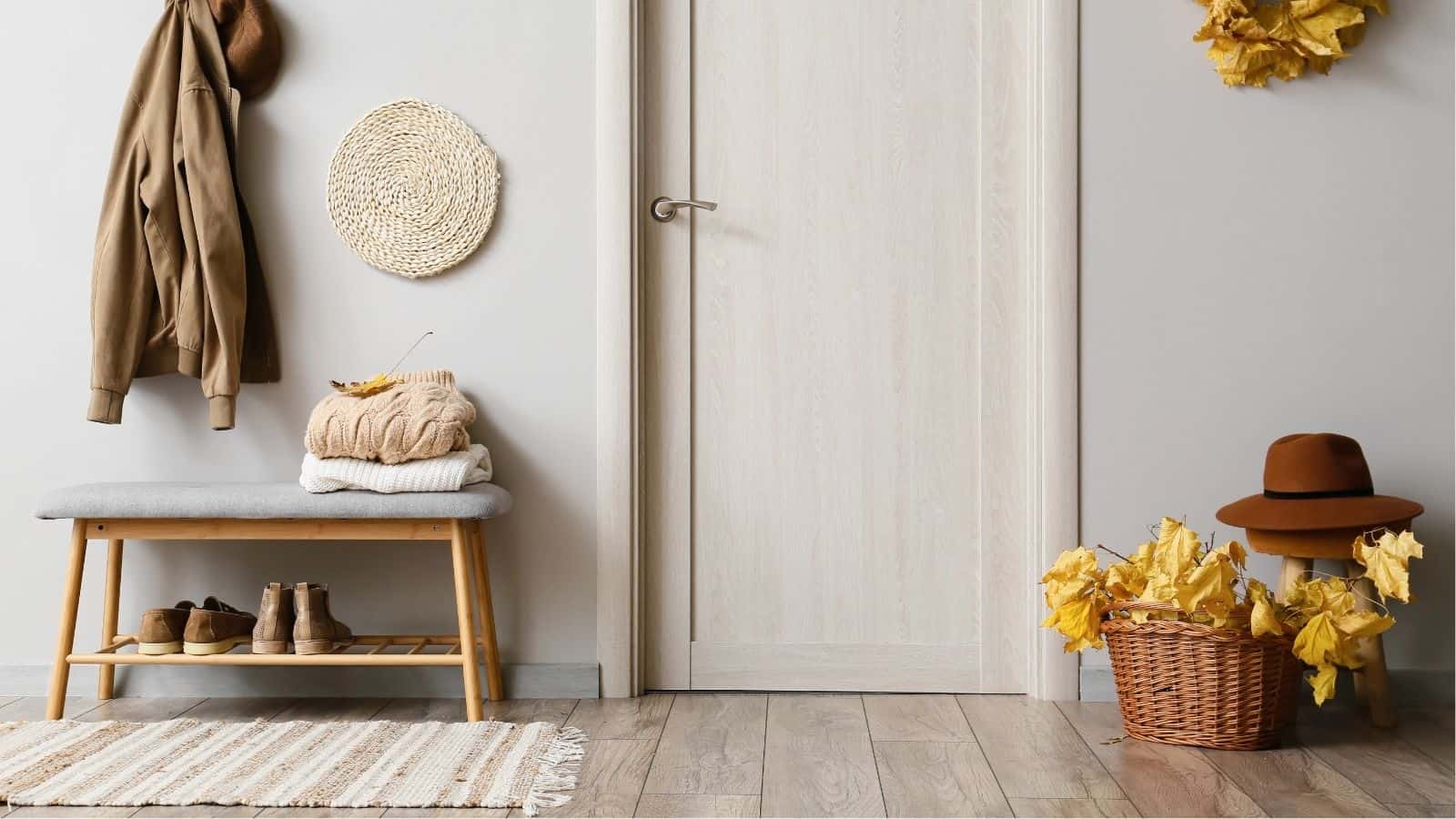 Neutral-toned entryway with a wooden bench, stacked sweaters, shoes, a jacket, hat, woven decor, a basket of yellow leaves, and a door in the background.