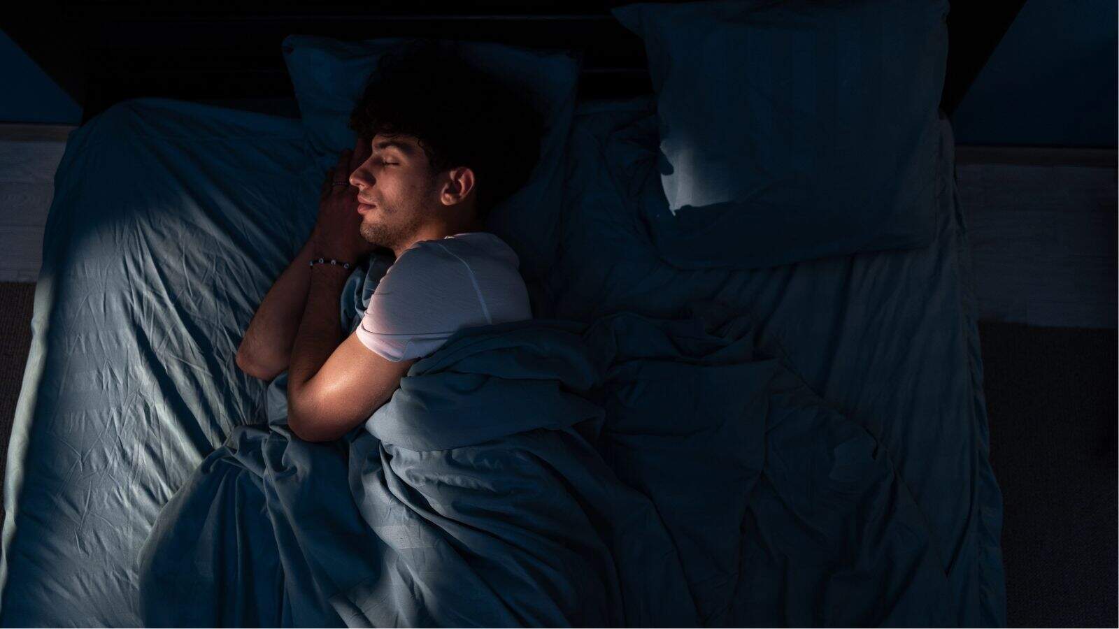 A man sleeps alone on a bed at night, lying on his side under a blue blanket with his head resting on a pillow.