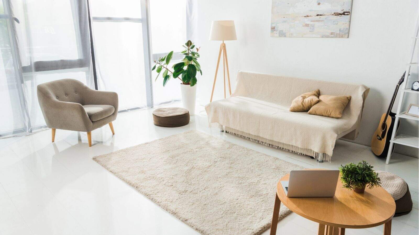 Bright living room with a beige sofa, armchair, floor lamp, houseplant, rug, and a small table holding a laptop and plant. A guitar leans against the wall near a window with sheer curtains.