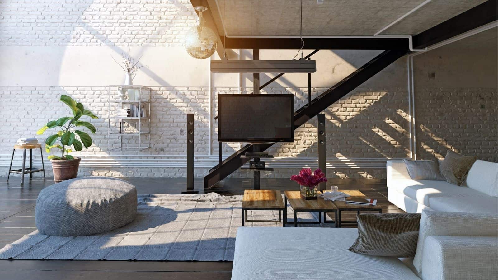 Modern loft living room with white brick walls, large windows, a white sectional sofa, a gray pouf, a flat-screen TV, and a staircase with a black metal railing.