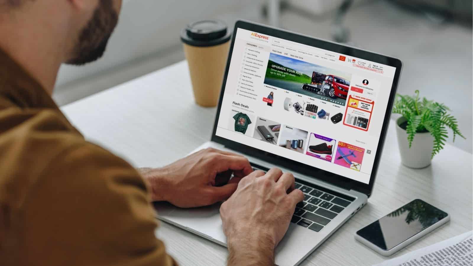 A person using a laptop browses an online shopping website displaying various products; a coffee cup and a small plant are on the desk.